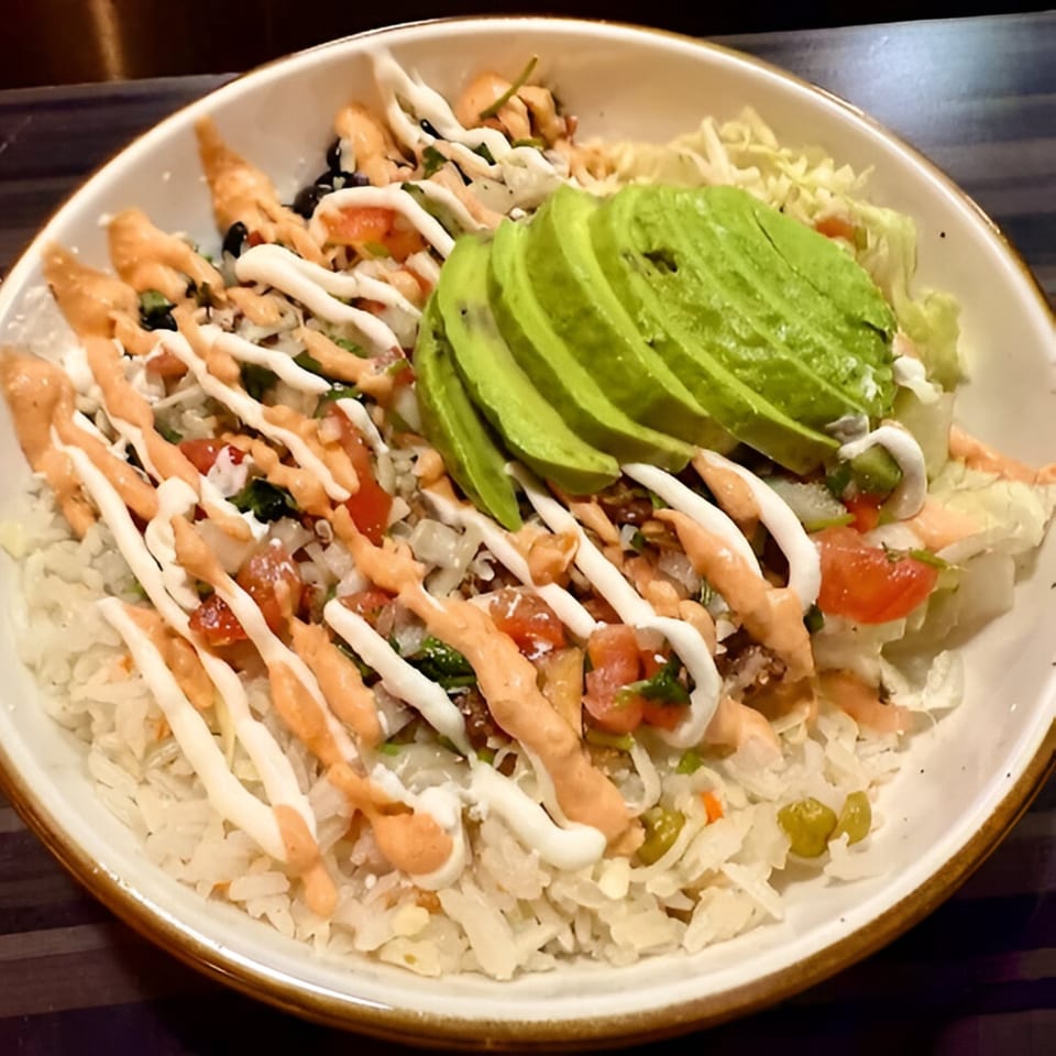 A Mexican rice bowl with sliced avocado, pico de gallo, and two sauces drizzled on top.